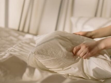 Hands arranging a pillow on a bed with light beige linens, creating a cozy bedroom ambiance.